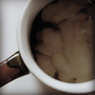 Photo of a coffee mug with cream beginning to swirl into the black coffee. The image shows only a portion of the cup looking down from above.